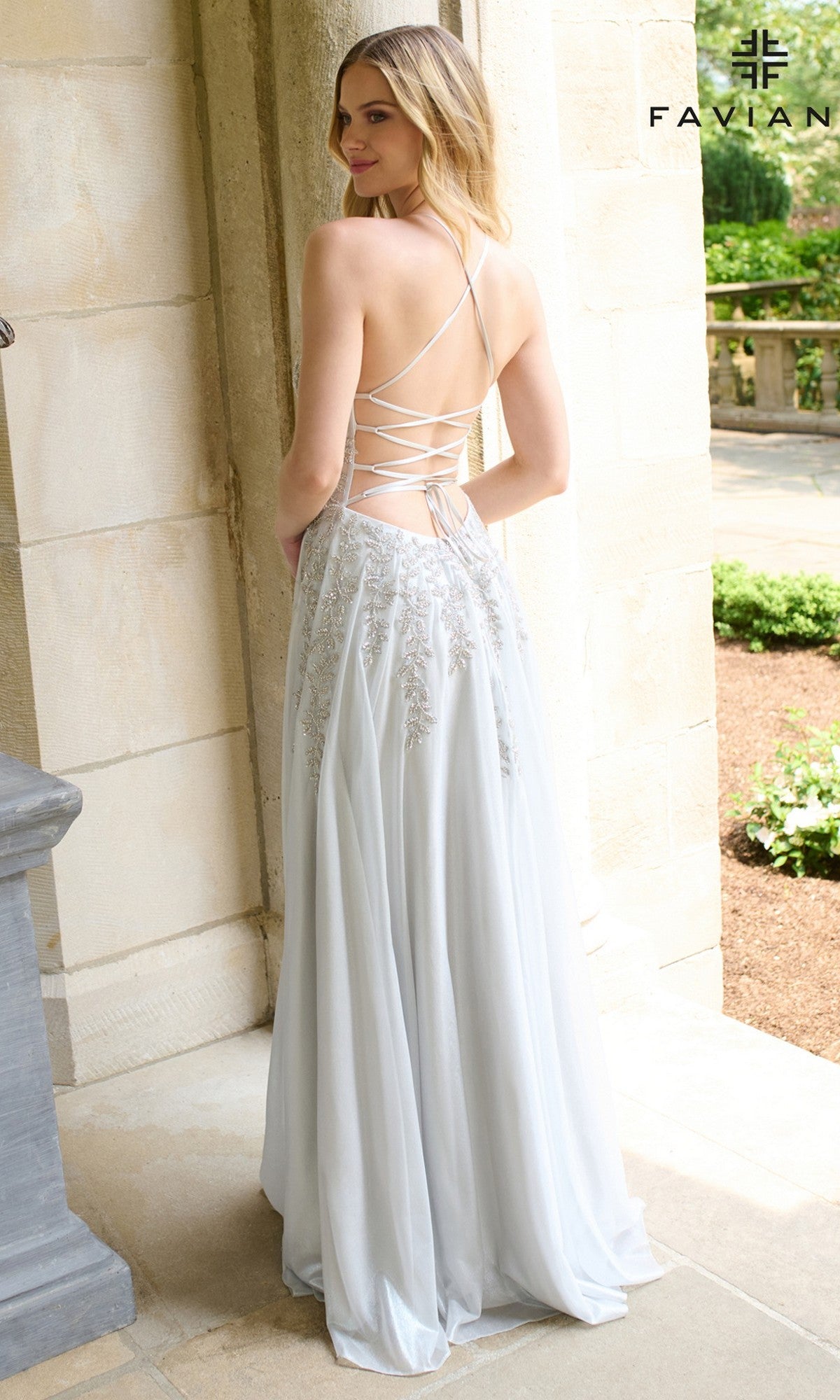 A woman stands outdoors, facing away from the camera, wearing the Long Prom Dress: Faviana 11449 in light blue with lace-up straps and embroidered details. The scene features stone columns and lush greenery.